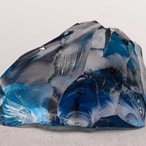 Puede incluir: Un gran trozo de vidrio azul transparente de forma irregular. El vidrio tiene una superficie rugosa y texturizada con bordes y facetas afiladas, que capturan la luz y crean reflejos y sombras. El objeto se muestra sobre una superficie gris neutra.