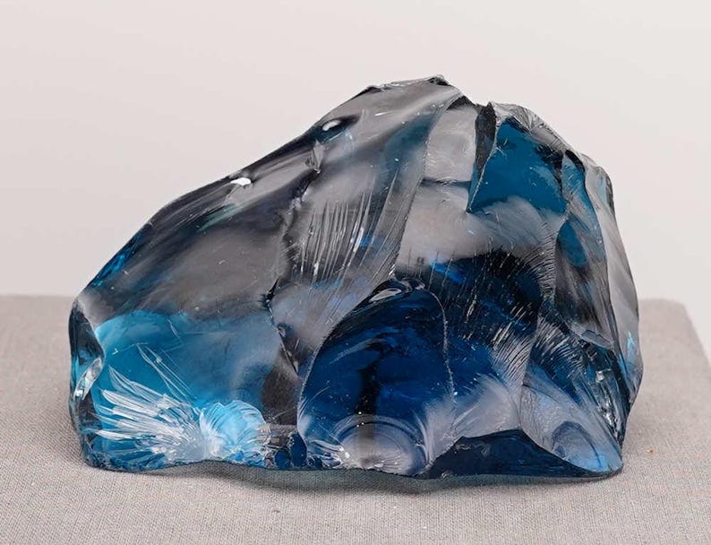 Puede incluir: Un gran trozo de vidrio azul transparente de forma irregular. El vidrio tiene una superficie rugosa y texturizada con bordes y facetas afiladas, que capturan la luz y crean reflejos y sombras. El objeto se muestra sobre una superficie gris neutra.