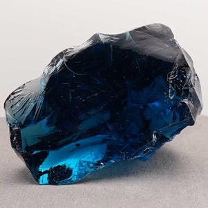 Puede incluir: Un gran trozo de vidrio azul oscuro de forma irregular. El vidrio tiene una superficie brillante y reflectante con bordes afilados y fracturados. El color var&iacute;a de un azul oscuro, casi negro, a un azul brillante y transl&uacute;cido. Objeto decorativo.