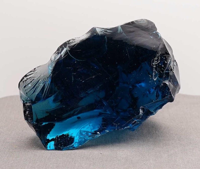 Puede incluir: Un gran trozo de vidrio azul oscuro de forma irregular. El vidrio tiene una superficie brillante y reflectante con bordes afilados y fracturados. El color var&iacute;a de un azul oscuro, casi negro, a un azul brillante y transl&uacute;cido. Objeto decorativo.