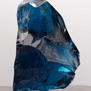 Puede incluir: Una pieza grande de vidrio azul de forma irregular, posiblemente un objeto decorativo o un esp&eacute;cimen mineral. El vidrio tiene una cualidad transl&uacute;cida con &aacute;reas de azul profundo y secciones m&aacute;s claras, casi transparentes. Se encuentra sobre una base cuadrada transparente.