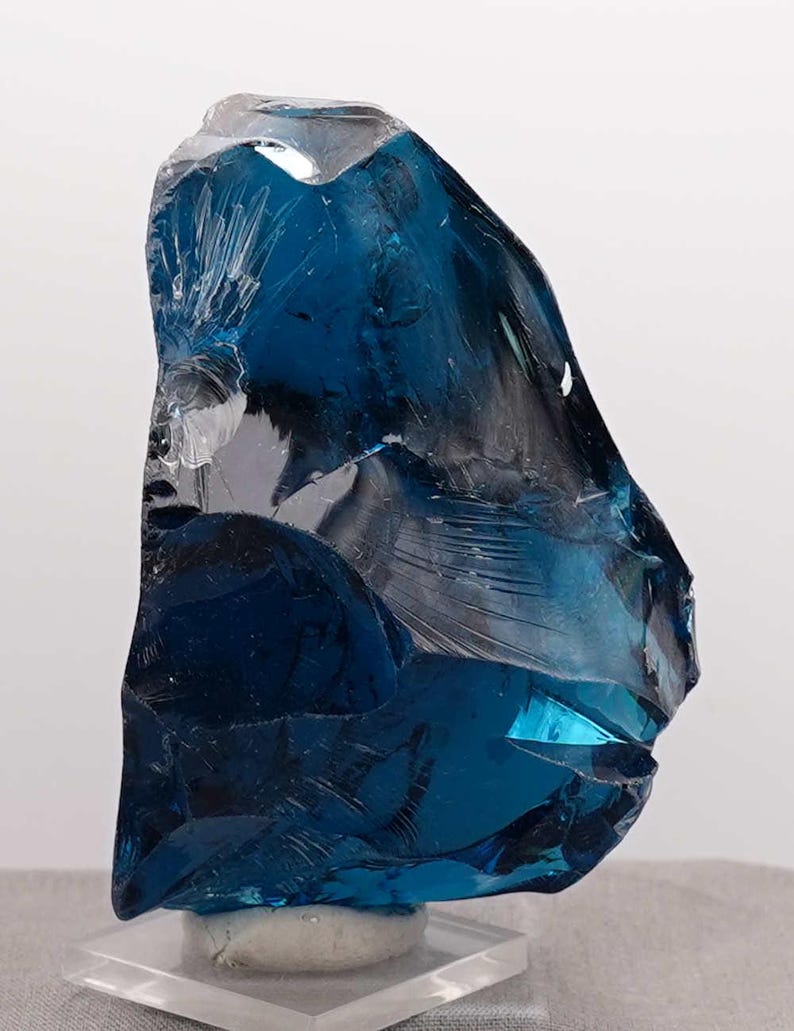 Puede incluir: Una pieza grande de vidrio azul de forma irregular, posiblemente un objeto decorativo o un esp&eacute;cimen mineral. El vidrio tiene una cualidad transl&uacute;cida con &aacute;reas de azul profundo y secciones m&aacute;s claras, casi transparentes. Se encuentra sobre una base cuadrada transparente.