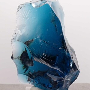 Puede incluir: Una gran pieza de vidrio azul de forma irregular, posiblemente una escultura decorativa. El vidrio tiene una calidad transl&uacute;cida con diferentes tonos de azul y blanco. Se asienta sobre una base rectangular transparente.