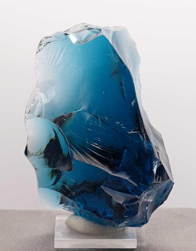 Puede incluir: Una gran pieza de vidrio azul de forma irregular, posiblemente una escultura decorativa. El vidrio tiene una calidad transl&uacute;cida con diferentes tonos de azul y blanco. Se asienta sobre una base rectangular transparente.