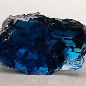 Puede incluir: Un gran trozo de vidrio azul oscuro de forma irregular. El vidrio tiene una superficie brillante y reflectante con fracturas internas y variaciones de color, creando una apariencia din&aacute;mica y texturizada. Probablemente una pieza decorativa o un objeto de arte.