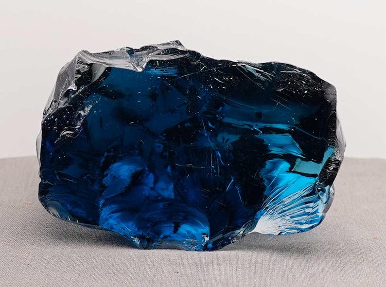 Puede incluir: Un gran trozo de vidrio azul oscuro de forma irregular. El vidrio tiene una superficie brillante y reflectante con fracturas internas y variaciones de color, creando una apariencia din&aacute;mica y texturizada. Probablemente una pieza decorativa o un objeto de arte.