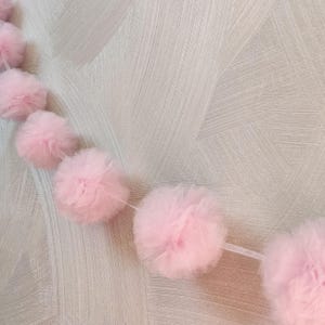 May include: A string of soft pink tulle pom-poms, strung together on a thin, clear thread. The pom-poms are evenly spaced and fluffy, creating a delicate and whimsical garland. The background is a textured, light-colored wall.