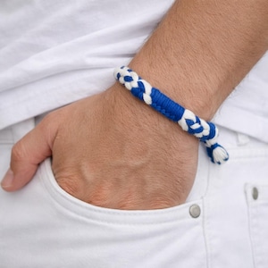 Puede incluir: Una pulsera trenzada en azul y blanco. La pulsera está hecha de un material grueso y tejido. La pulsera se lleva en la muñeca, con la mano en un bolsillo. El fondo es una camisa blanca y pantalones blancos.