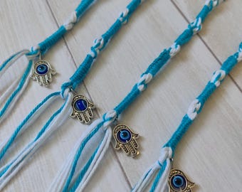 May include: Close-up of several blue and white braided bracelets. Each bracelet features a silver charm in the shape of a hand with a blue eye in the center. The bracelets are made of braided cord.