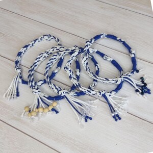Op de afbeelding: Gevlochten blauw-witte armbanden. Elke armband is gemaakt van geweven koord en heeft een mix van witte en blauwe draden. Sommige hebben houten kralen of kleine, donkerblauwe kralen.