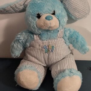 Peut inclure: Un lapin en peluche bleu avec un visage blanc et des yeux bleus. Le lapin porte une salopette rayée bleu et blanc avec le texte "Baby Stuff" sur le devant.