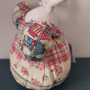May include: A fabric bunny doorstop with a white body and patterned ears and clothing. The ears have a pink and green plaid design, and the body features floral and patchwork patterns. The doorstop is designed to stand upright.