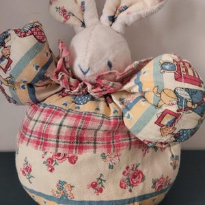 May include: A soft bunny doll with a white head and ears, and a round body. The body is covered in a floral fabric with red, pink, and yellow accents. The bunny has a pink ruffled collar.