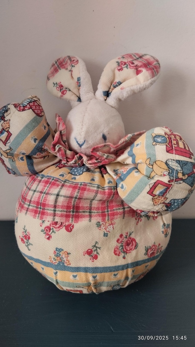 May include: A soft bunny doll with a white head and ears, and a round body. The body is covered in a floral fabric with red, pink, and yellow accents. The bunny has a pink ruffled collar.