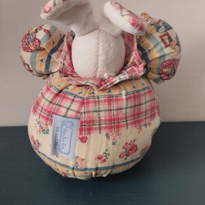May include: A plush bunny ornament with a white head and patterned ears. The body is round, covered in a plaid and floral fabric. A small label is attached to the body. The item is a decorative piece.