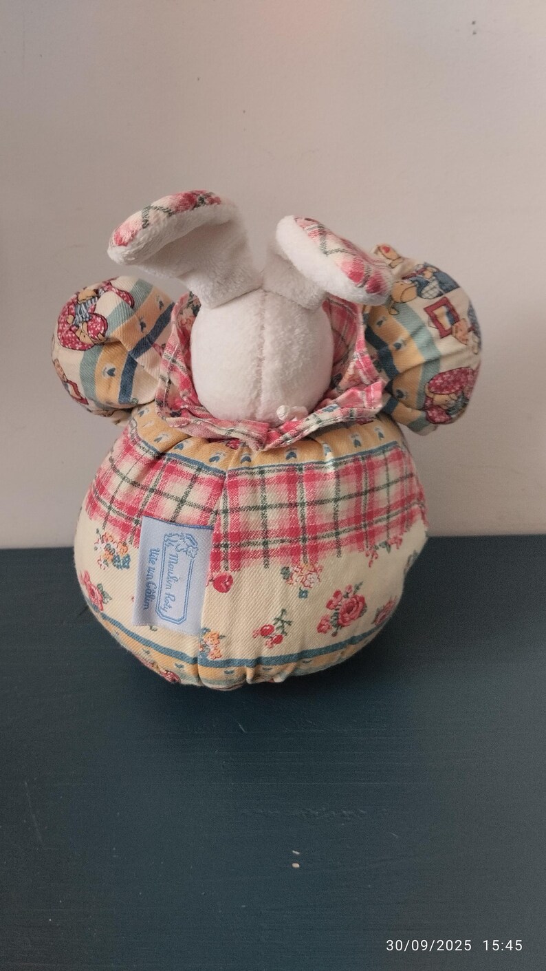 May include: A plush bunny ornament with a white head and patterned ears. The body is round, covered in a plaid and floral fabric. A small label is attached to the body. The item is a decorative piece.