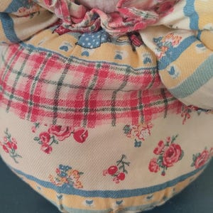 May include: A handmade doll with a white plush head and a floral patterned dress. The dress features a mix of red, yellow, and blue floral and plaid patterns. The doll has blue stitched eyes and a pink ruffled collar.