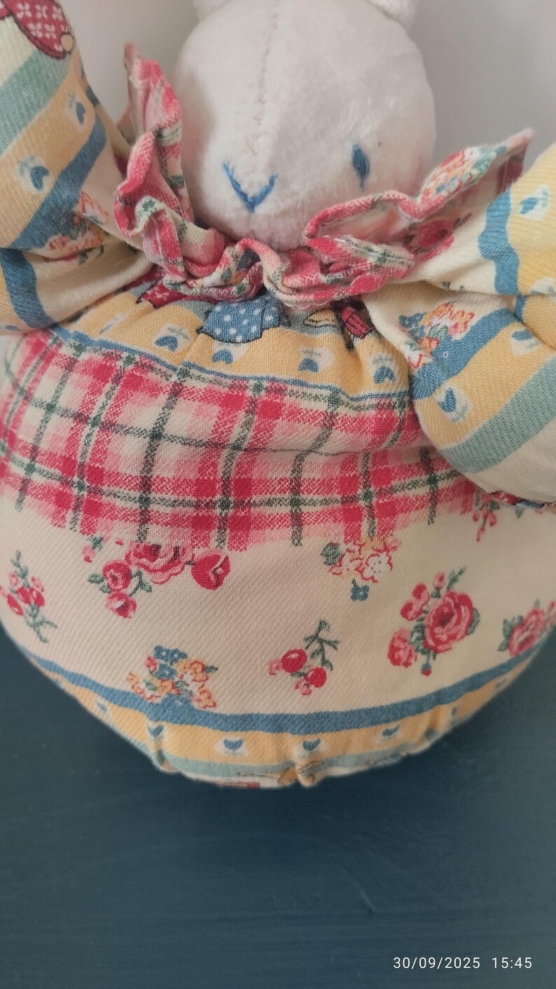 May include: A handmade doll with a white plush head and a floral patterned dress. The dress features a mix of red, yellow, and blue floral and plaid patterns. The doll has blue stitched eyes and a pink ruffled collar.
