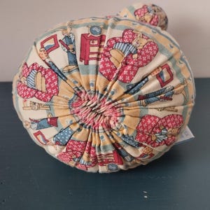 May include: A round, fabric-covered decorative pillow with a gathered center. The pillow features a repeating pattern of figures in red, blue, and yellow on a cream background. The pillow is approximately 15 cm in diameter.