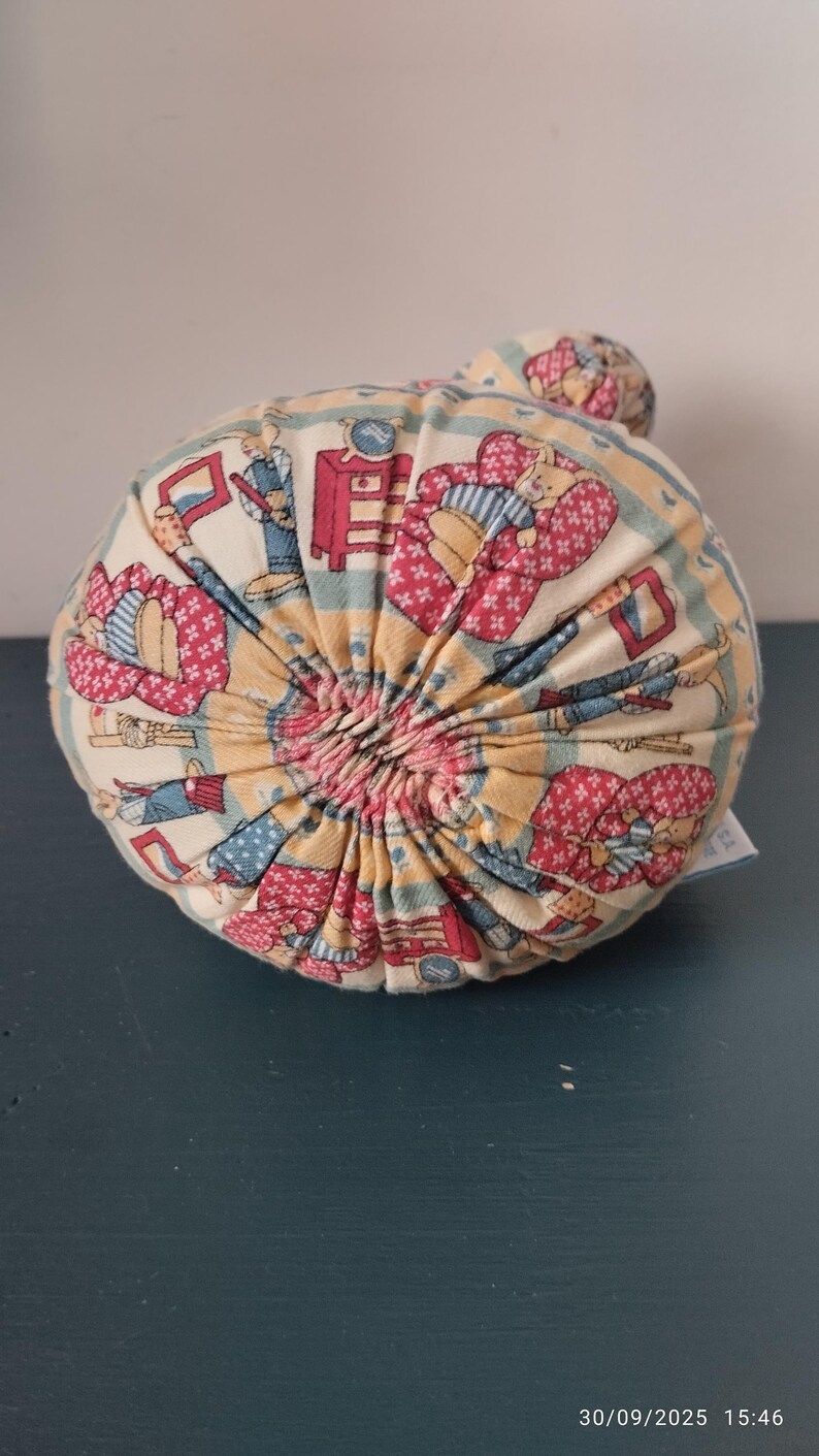 May include: A round, fabric-covered decorative pillow with a gathered center. The pillow features a repeating pattern of figures in red, blue, and yellow on a cream background. The pillow is approximately 15 cm in diameter.