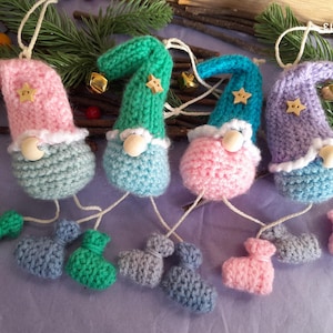May include: Four handmade crochet gnome decorations in pink, green, blue, and purple. Each gnome has a white beard, a star on its hat, and a small bell. The gnomes have dangling legs and are suspended by white string.