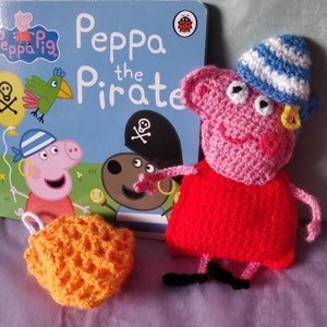 May include: A crocheted Peppa Pig doll dressed as a pirate. She is wearing a red dress, a blue and white striped hat, and a gold earring. The doll is holding a sword and is standing in front of a book titled "Peppa the Pirate".