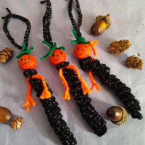 May include: Three handmade Halloween ornaments. Each ornament has an orange pumpkin head with a carved face, green stem, and black and orange body. Acorns and pinecones are scattered around the ornaments.