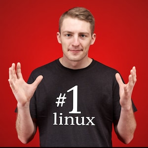 Peut inclure: Un t-shirt gris foncé avec le texte "#1 linux" en blanc. La personne sur l'image sourit et a les mains levées. L'arrière-plan est rouge uni.