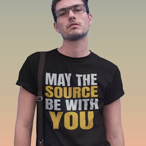Peut inclure: Un t-shirt noir avec le texte "Que la source soit avec toi" en lettres blanches et dorées.