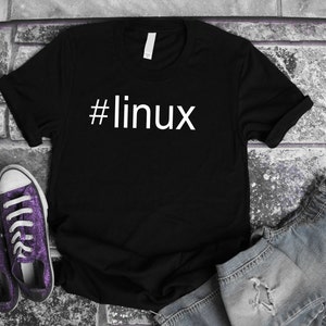 May include: Black t-shirt with the white text "#linux". The shirt is displayed with purple glitter sneakers and a pair of denim shorts on a brick surface. The t-shirt is a casual wear item.