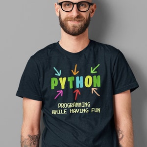May include: A dark blue t-shirt with the word "PYTHON" in colorful letters surrounded by colorful arrows. The text "PROGRAMMING WHILE HAVING FUN" is printed below the word "PYTHON" in a pixelated font.