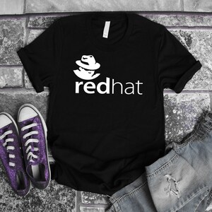 Peut inclure: T-shirt noir avec le logo "redhat" blanc et un graphisme d'une personne portant un chapeau. L'image présente également des baskets violettes à paillettes et un short en jean.