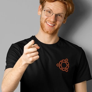 Peut inclure: Un homme aux cheveux roux et à la barbe porte un t-shirt noir avec un logo Linux orange. Il sourit et pointe du doigt vers le spectateur.
