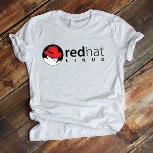 Peut inclure: T-shirt blanc avec le logo Red Hat Linux. Le logo comprend un chapeau rouge, un cercle noir et les mots "red hat LINUX" en noir. Le t-shirt est fait d'une matière douce.