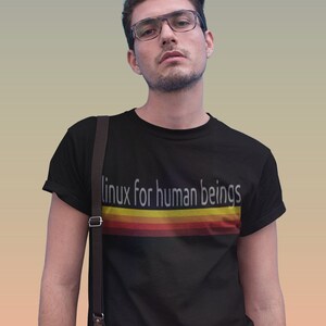 Peut inclure: Un t-shirt noir avec un graphisme rétro qui dit "linux for human beings" en texte blanc. Le graphisme présente une série de rayures colorées en rouge, orange, jaune et or.