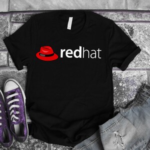 Peut inclure: T-shirt noir avec le texte blanc "redhat" et un chapeau fedora rouge. Le t-shirt est présenté sur une surface en briques grises, avec des baskets violettes et un short en jean en arrière-plan.