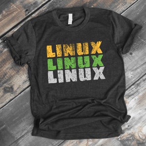 May include: Dark gray t-shirt with the word "LINUX" printed three times in yellow, green, and white. The text has a distressed, vintage look. The shirt is made of a soft material and has a crew neck.