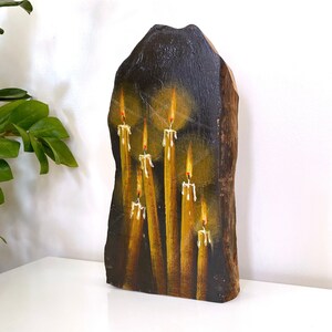 May include: A hand-painted wooden plaque featuring a candle design. The artwork depicts several yellow candles with orange flames and white wax drips against a dark brown background. The wooden plaque has a natural, irregular shape.