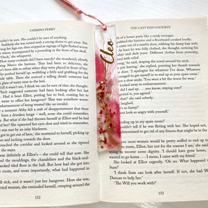 White and pink resin bookmark with dried roses and gold leaf, personalised with gold vinyl, placed inside an open book, handmade bookmark for readers or gifts.