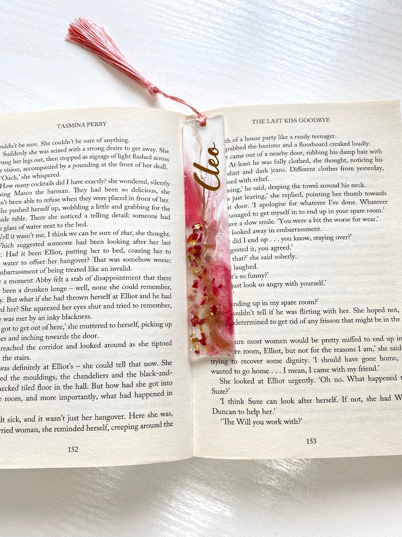 White and pink resin bookmark with dried roses and gold leaf, personalised with gold vinyl, placed inside an open book, handmade bookmark for readers or gifts.