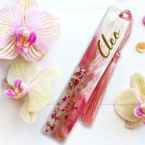 K&ouml;nnte beinhalten: Ein rosa-wei&szlig;es Lesezeichen aus Harz mit goldenen Flocken und einem rosa Quasten. Das Lesezeichen tr&auml;gt den Namen "Cleo" in goldenen Buchstaben.