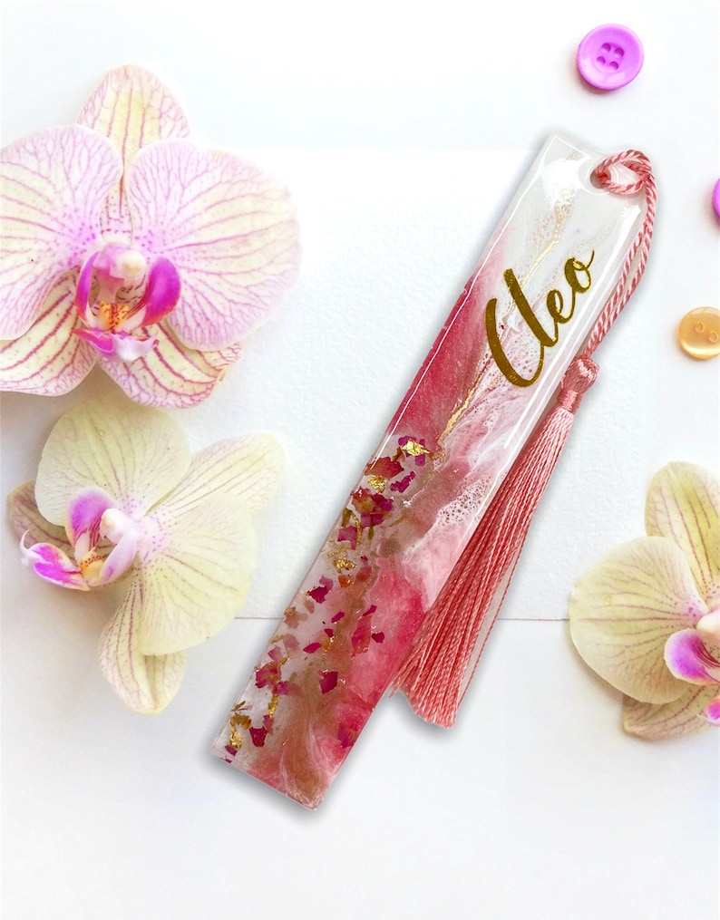 K&ouml;nnte beinhalten: Ein rosa-wei&szlig;es Lesezeichen aus Harz mit goldenen Flocken und einem rosa Quasten. Das Lesezeichen tr&auml;gt den Namen "Cleo" in goldenen Buchstaben.