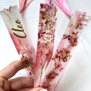 Hand holding three white and pink resin bookmarks with dried roses and gold leaf, including personalised and non-personalised options, handmade bookmarks for readers or gifts