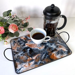 May include: A black and white marble tray with a black metal frame. A white ceramic coffee cup with coffee and a black French press coffee maker are on the tray.