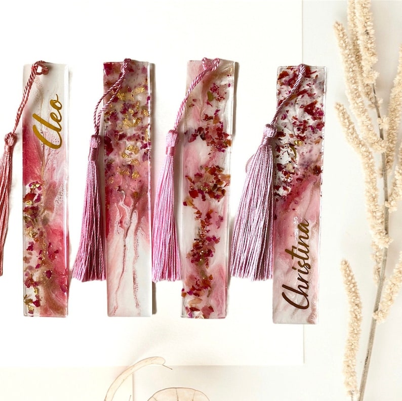 K&ouml;nnte beinhalten: Vier personalisierte Lesezeichen aus rosa und goldenem Harz, mit getrockneten Blumen und rosa Quasten. Die Lesezeichen sind mit "Cleo" und "Christina" beschriftet.