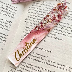 White and pink resin bookmark with dried roses and gold leaf, personalised with gold vinyl, placed inside an open book, handmade bookmark for readers or gifts.