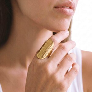May include: A gold-toned ring featuring a large, elongated, oval-shaped face with a distinct hammered, dimpled texture. The metal surface has a matte finish, covering the upper portion of the finger with a wide, curved, and solid band design.