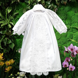 May include: A white baby dress with lace detailing. The dress features a round neckline and three-quarter sleeves with lace trim. A vertical panel of intricate lace adorns the front of the dress, adding a delicate touch. The dress is hanging on a white hanger.