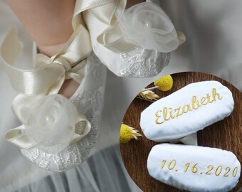 Könnte beinhalten: Weiße Satin-Babyschuhe mit Spitzendetails und einer weißen Satinschleife. Die Schuhe sind mit einer weißen Tüllblume und einer Perle verziert. Die Schuhe sind auch mit dem Namen "Elizabeth" und dem Datum "10.16.2020" in goldener Stickerei personalisiert.