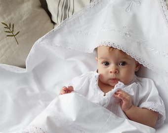 Könnte beinhalten: Eine weiße Babydecke mit Spitzenbesatz und gestickten Blumen. Ein Baby ist in die Decke gewickelt und trägt ein weißes Taufkleid mit Spitzenbesatz.